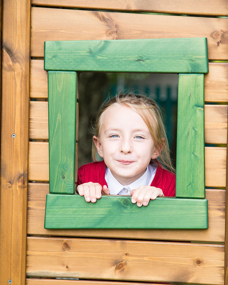 Ein kleines Mädchen schaut durch ein Holzfenster eines Spielhauses auf einem Spielplatz und lächelt.